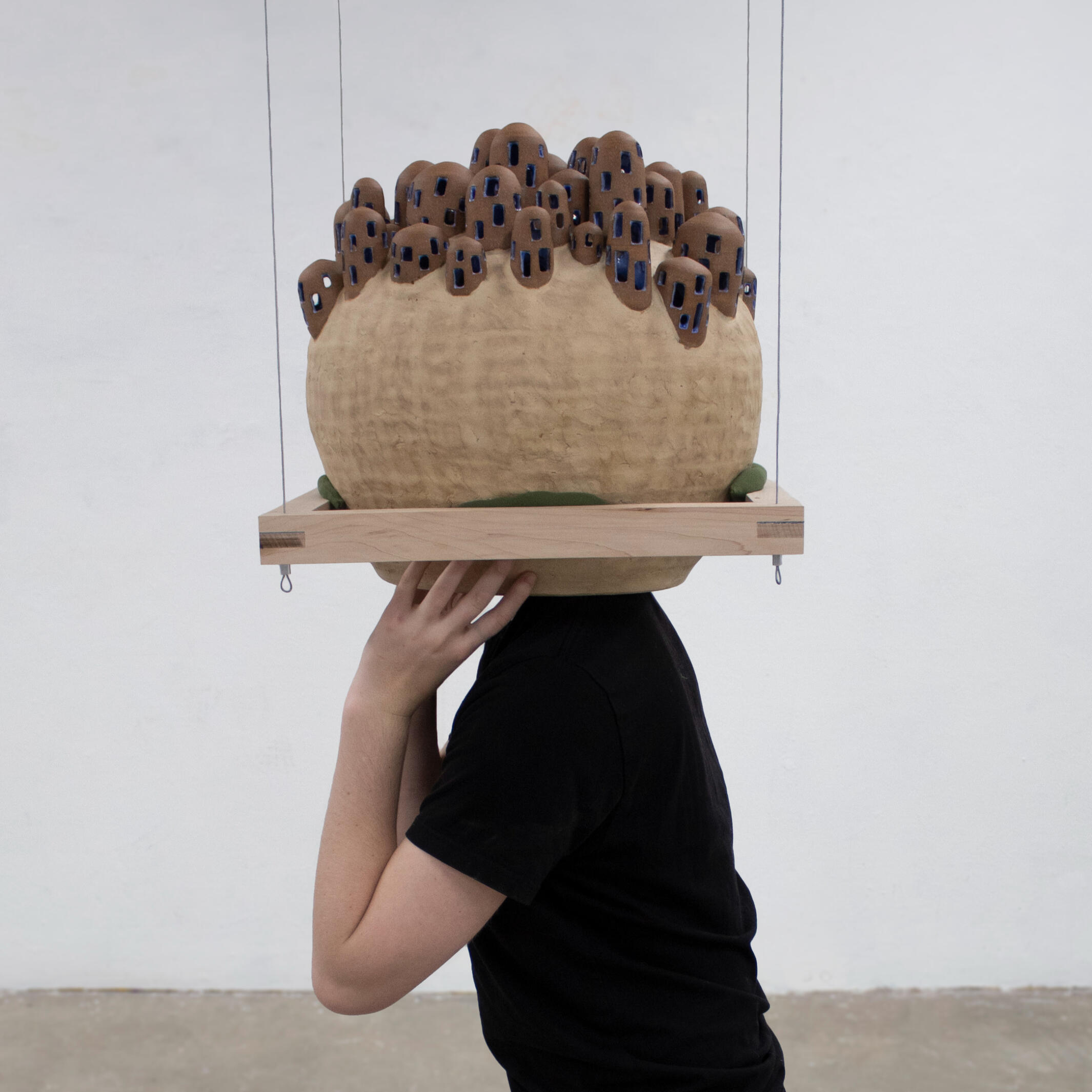 A person in black clothing has their head stuck in a handcrafted ceramic sphere suspended by a wooden frame and wire cables against a white background. The sphere has 50 small round houses made of clay on top of it. Made by Mia Donlason.