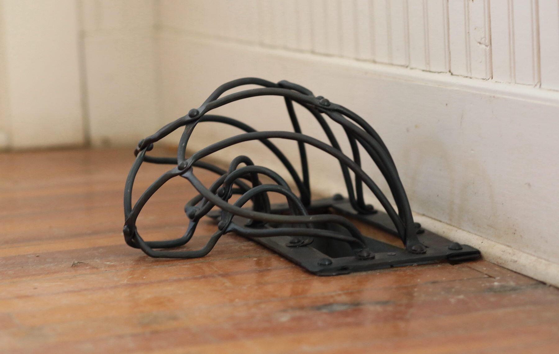 A handcrafted forged and fabricated steel grate installed covering a wooden floor vent hole with white trim on the wall behind. Made by Mia Donalson.