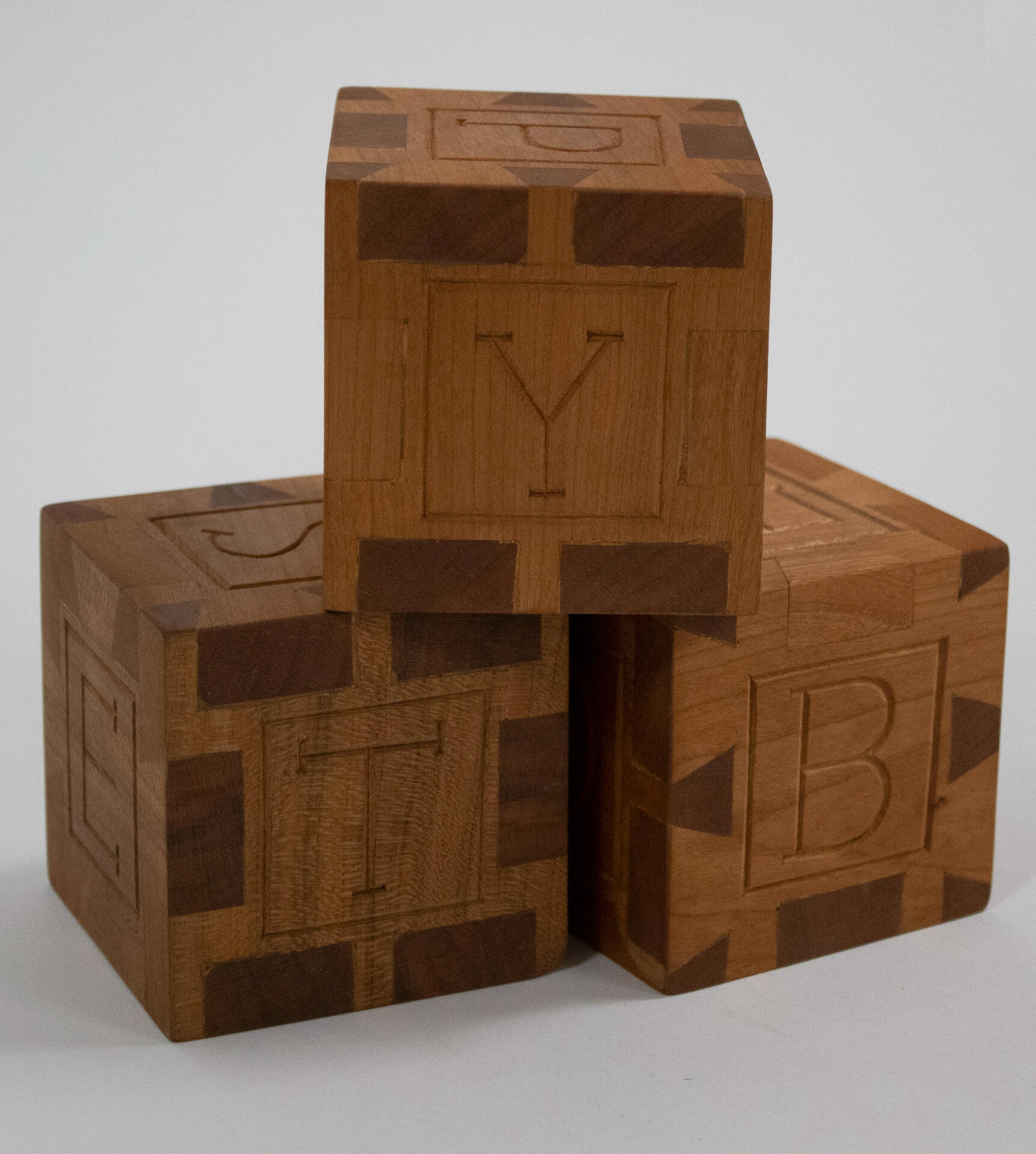 Three hand carved cherry wood building blocks sit stacked in a triangle against a white background. They are constructed using a handcut dovetail joint and have one capital english letter carved on each face of the cube. Made by Mia Donalson.