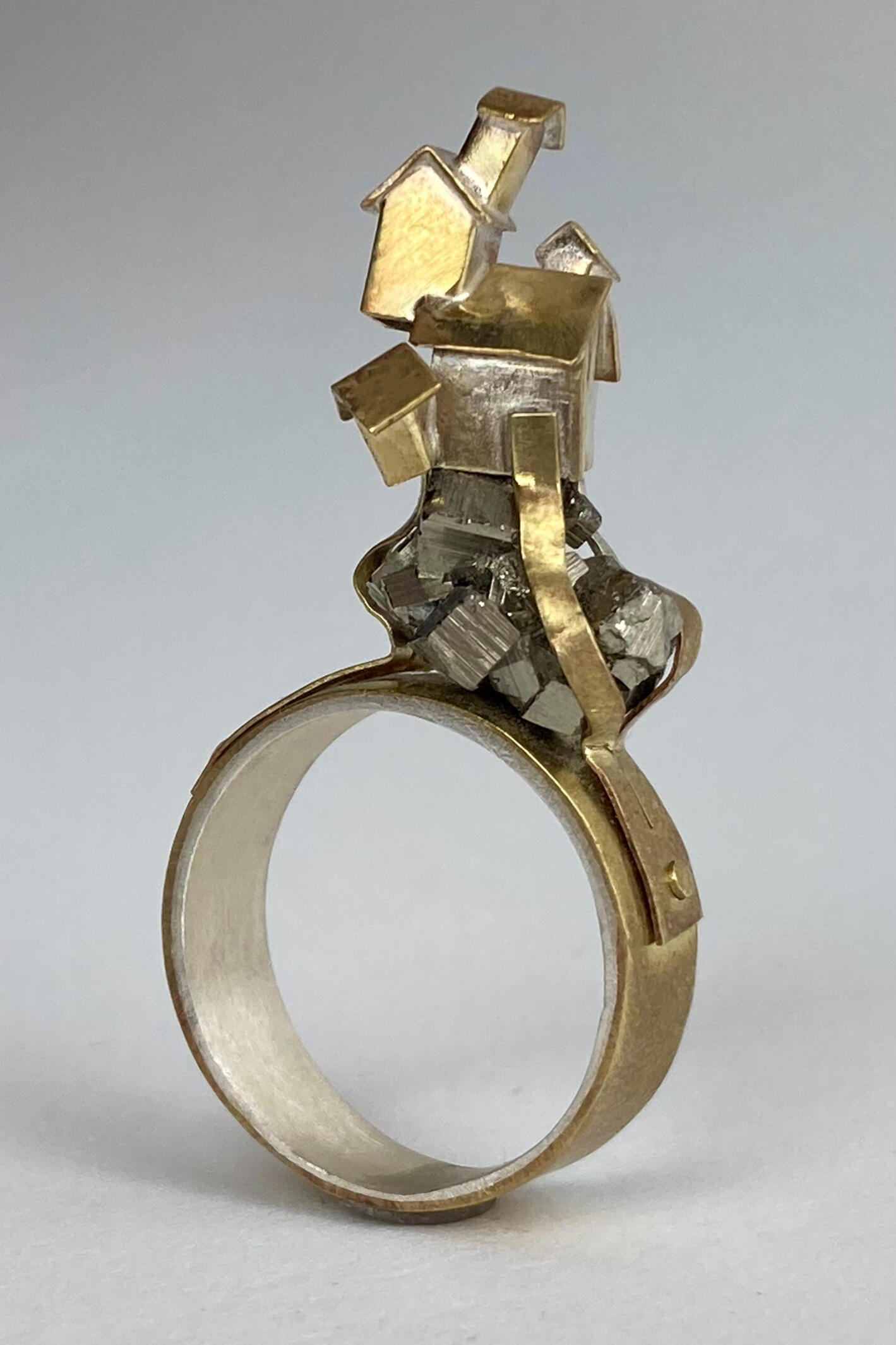 A handcrafted brass ring with a silver lining is shown against a white background. There is a piece of pyrite stone set with brass bands and small fabricated houses on the top of the ring. Made by Mia Donalson.