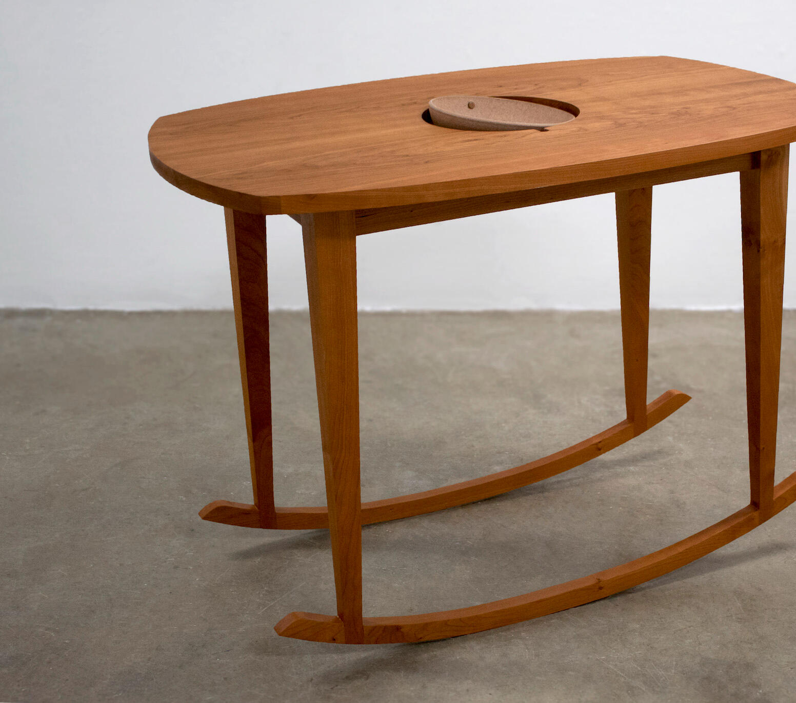 A handcrafted cherry wood table with rockers sits on a concrete floor with a white wall background. The table has a ceramic bowl inset into the center of the table top like a gyroscope. Made my Mia Donalson.