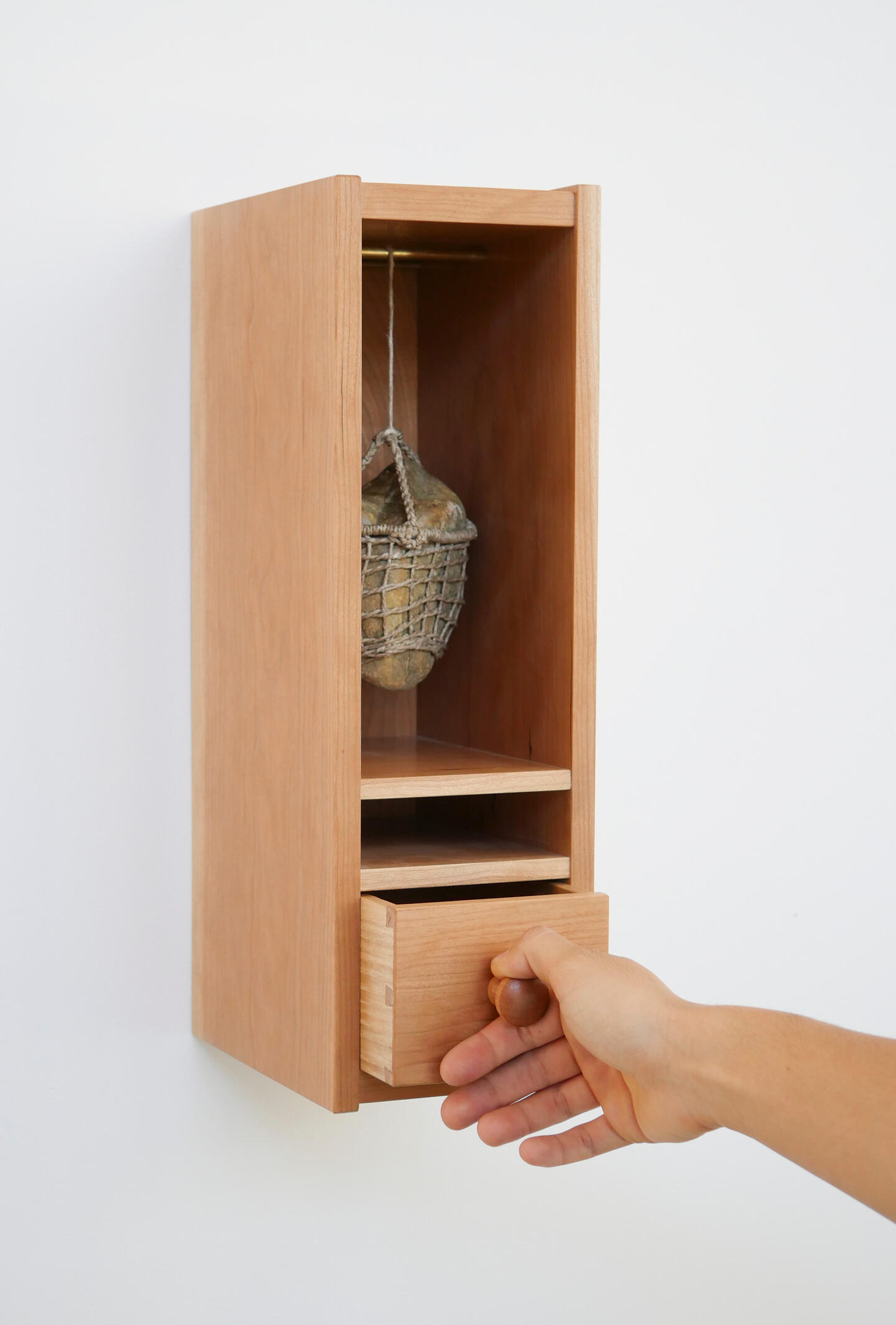 A small cherry wood cabinet hung on a white wall has an open shelf in the top two thirds and a drawer in the bottom one third. The drawer is being partially opened by a hand and that action is lifting a rock on a cable in the upper cavity. Made by Mia Dona
