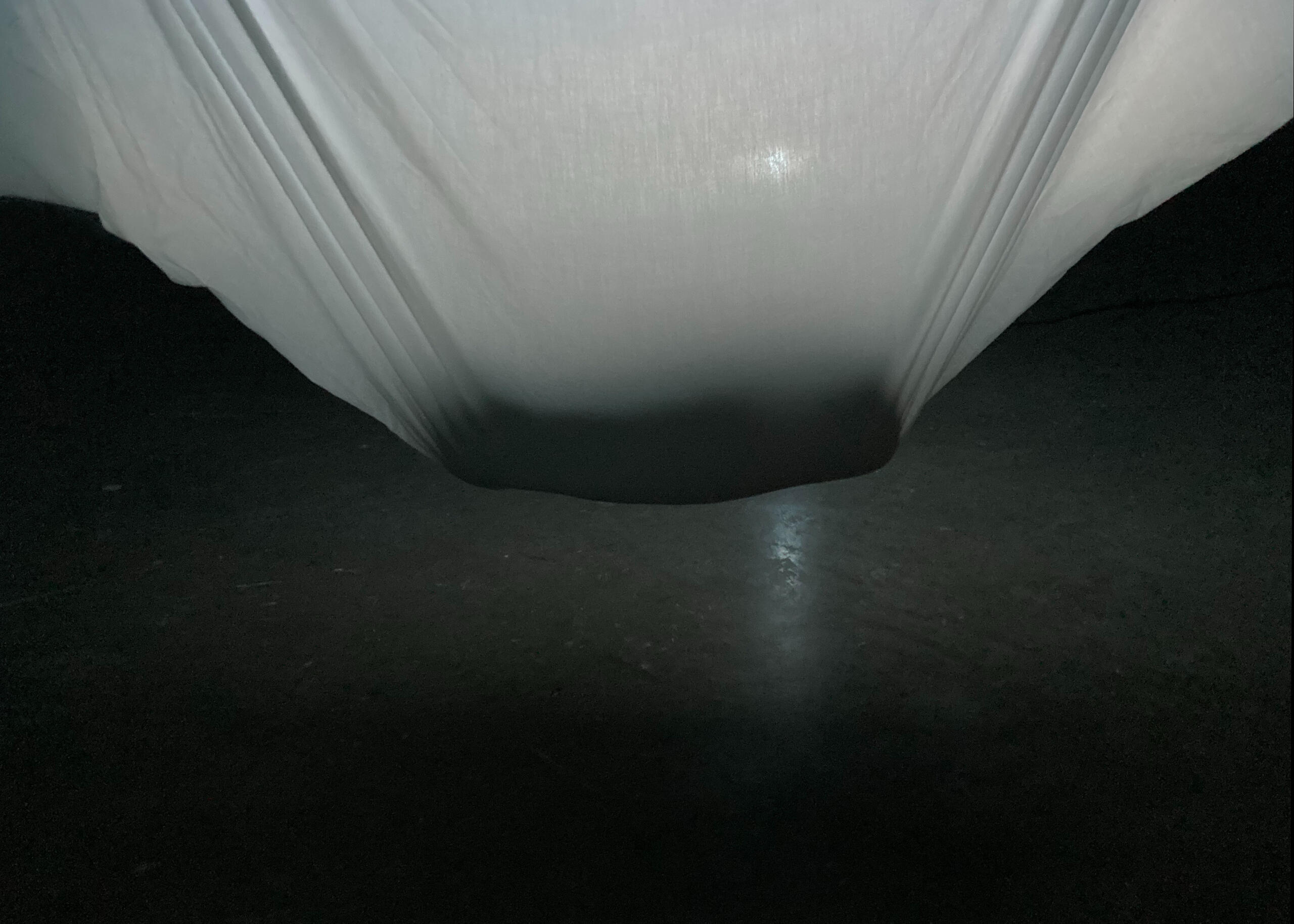 A white sheet hangs backlight with a ceramic object suspended above a concrete floor. Made by Mia Donalson.