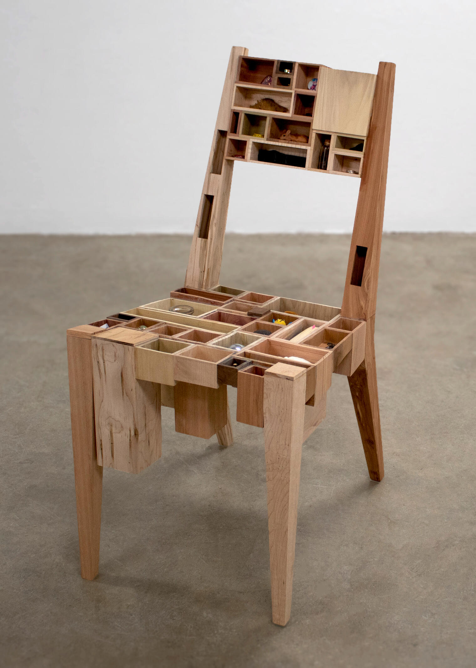A handcrafted wooden chair sits on a concrete floor with a white wall background. 50 boxes of different wood and size make up the shape of the chair and each box holds a different object of importance. Made by Mia Donalson.