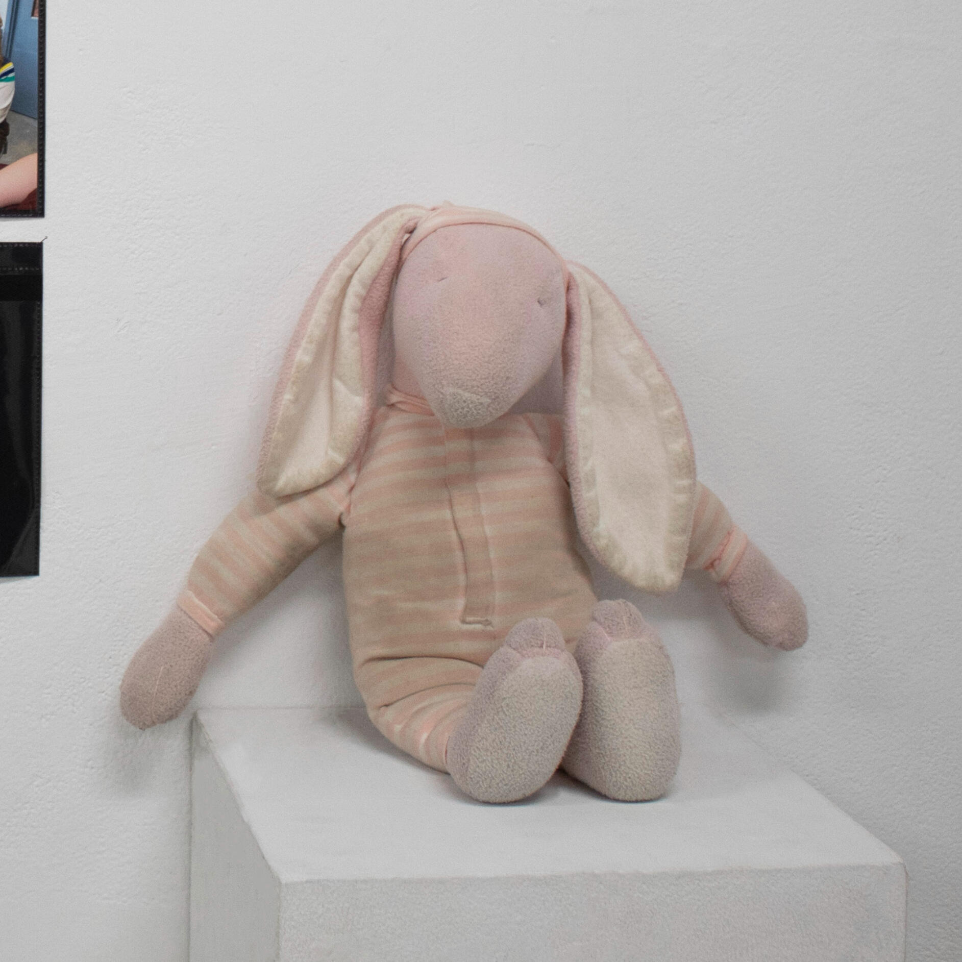 A hand sewn pink and white striped stuffed animal rabbit with a hat and some dirt on it sitting on a white pedestal against a white wall. Made by Mia Donalson.