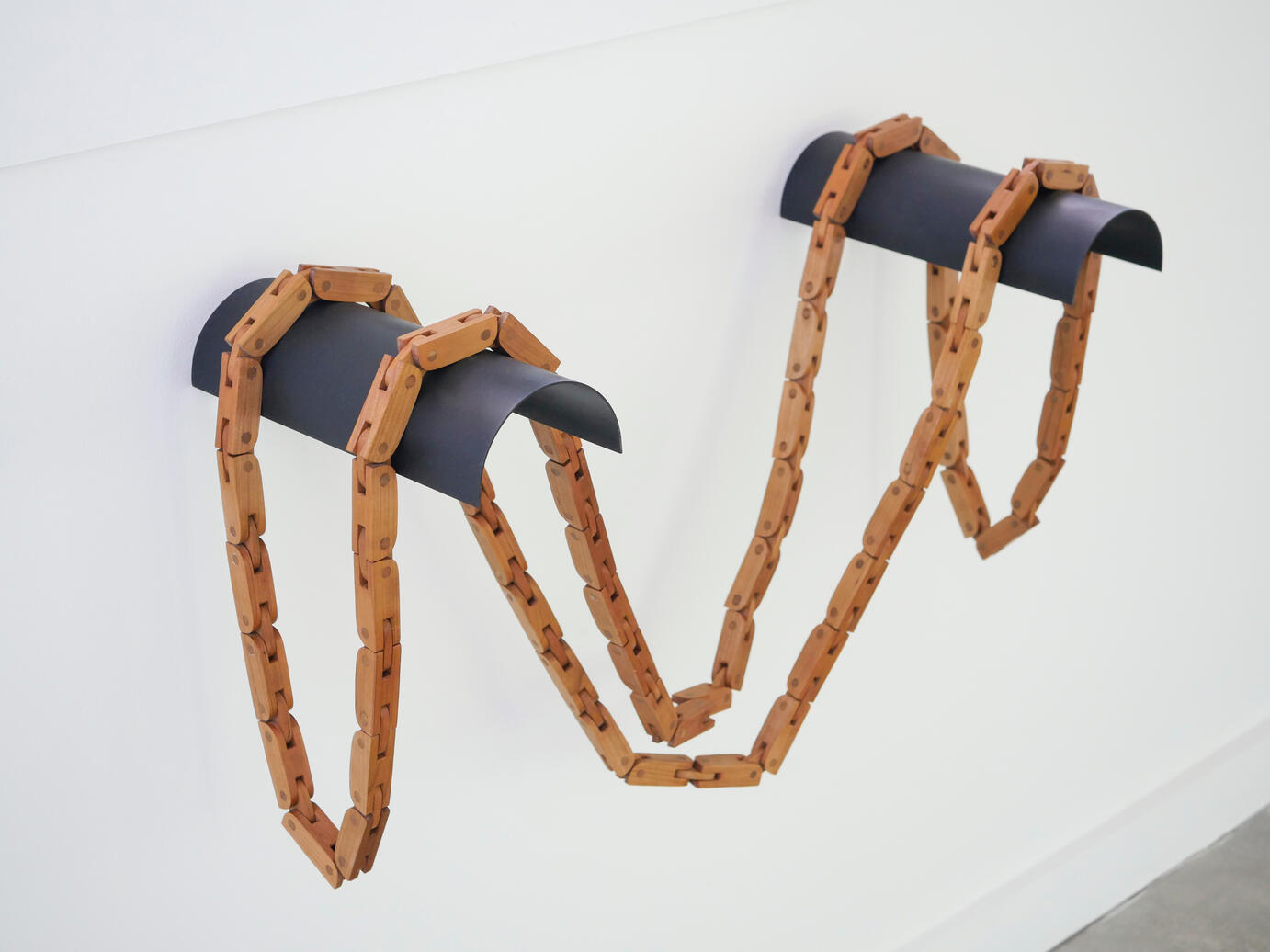 Two people stand facing each other wearing the artwork called Softened. Softened is a handcrafted cherry wood articulated chain. A handcrafted table made from cherry wood with an orange-brown cotton fabric top shown against a white background. It is held up from collape by two human hands. Made by Mia Donalson.