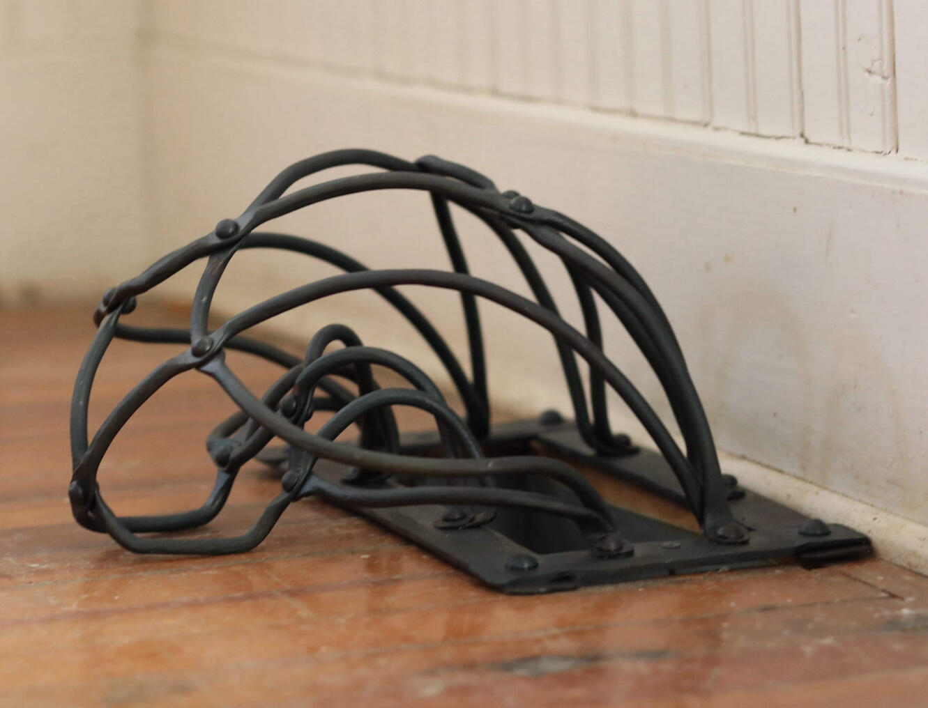 A handcrafted forged and fabricated steel grate installed covering a wooden floor vent hole with white trim on the wall behind. Made by Mia Donalson.