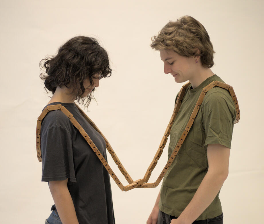 Two people stand facing each other wearing the artwork called Softened. Softened is a handcrafted cherry wood articulated chain. A handcrafted table made from cherry wood with an orange-brown cotton fabric top shown against a white background. It is held up from collape by two human hands. Made by Mia Donalson.