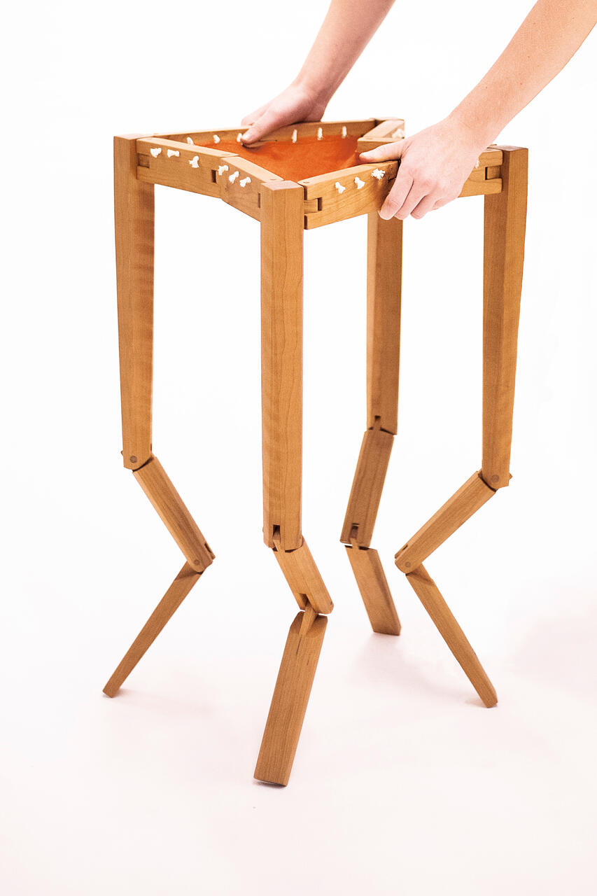 A handcrafted table made from cherry wood with an orange-brown cotton fabric top shown against a white background. It is held up mid-collape by two human hands. Made by Mia Donalson.