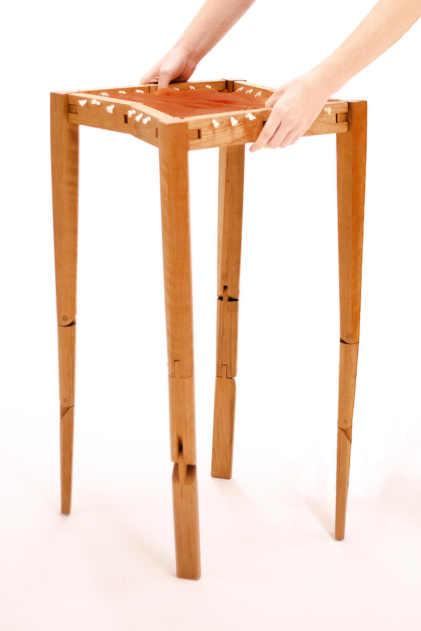 A handcrafted table made from cherry wood with an orange-brown cotton fabric top shown against a white background. It is held up from collape by two human hands. Made by Mia Donalson.