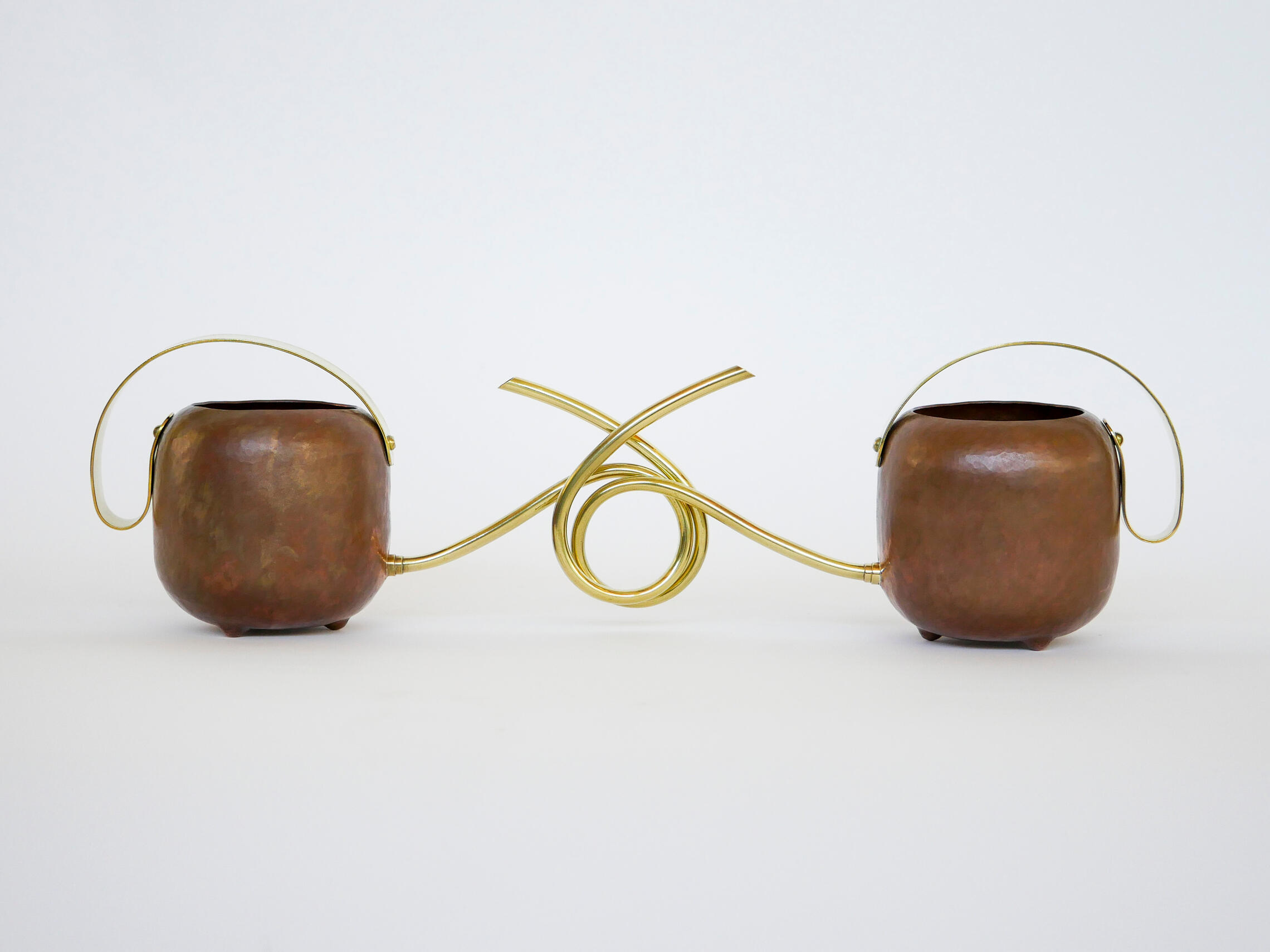 A pair of handcrafted watering cans made from brass and copper is shown against a white background. The spouts of each are coiled to lock around each other and are connected. Made by Mia Donalson.