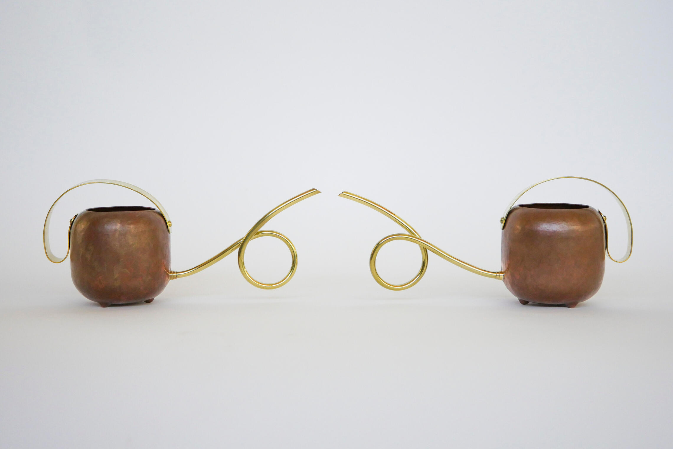 A pair of handcrafted watering cans made from brass and copper is shown against a white background. The spouts of each are coiled to lock around each other but are pictured separate and facing. Made by Mia Donalson.