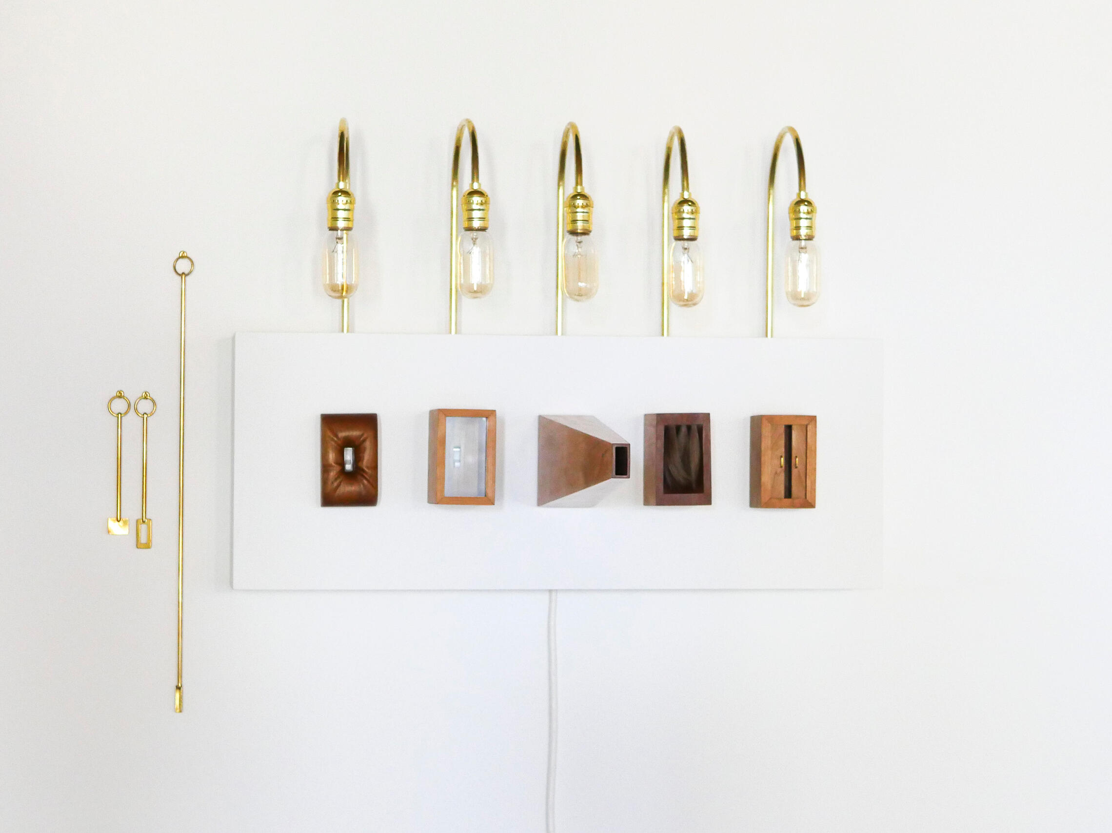 Five unique light switch covers are mounted to a white box on the wall. Five light bulbs on bent brass tube stick out of the top of the box. There are three brass tools hung on the wall left of the box. Made by Mia Donalson.