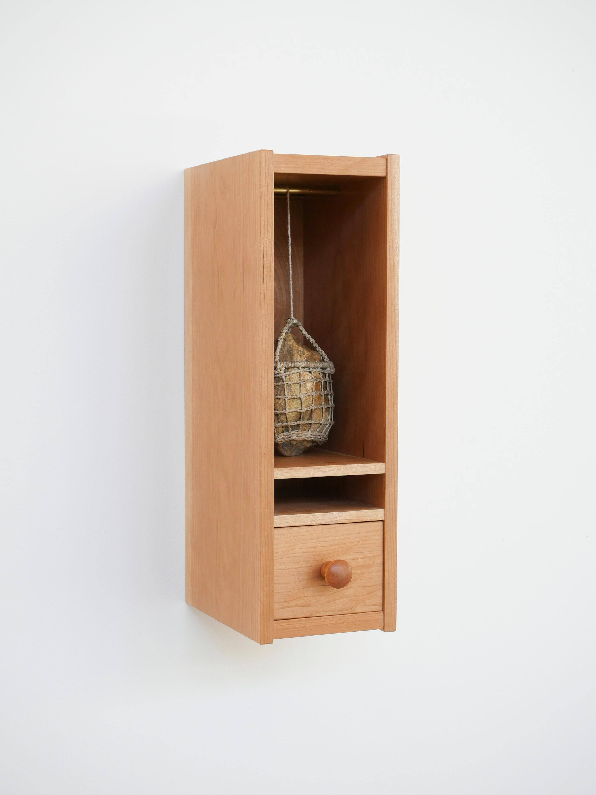 A small cherry wood cabinet hung on a white wall has an open shelf in the top two thirds and a drawer in the bottom one third. Made by Mia Donalson.