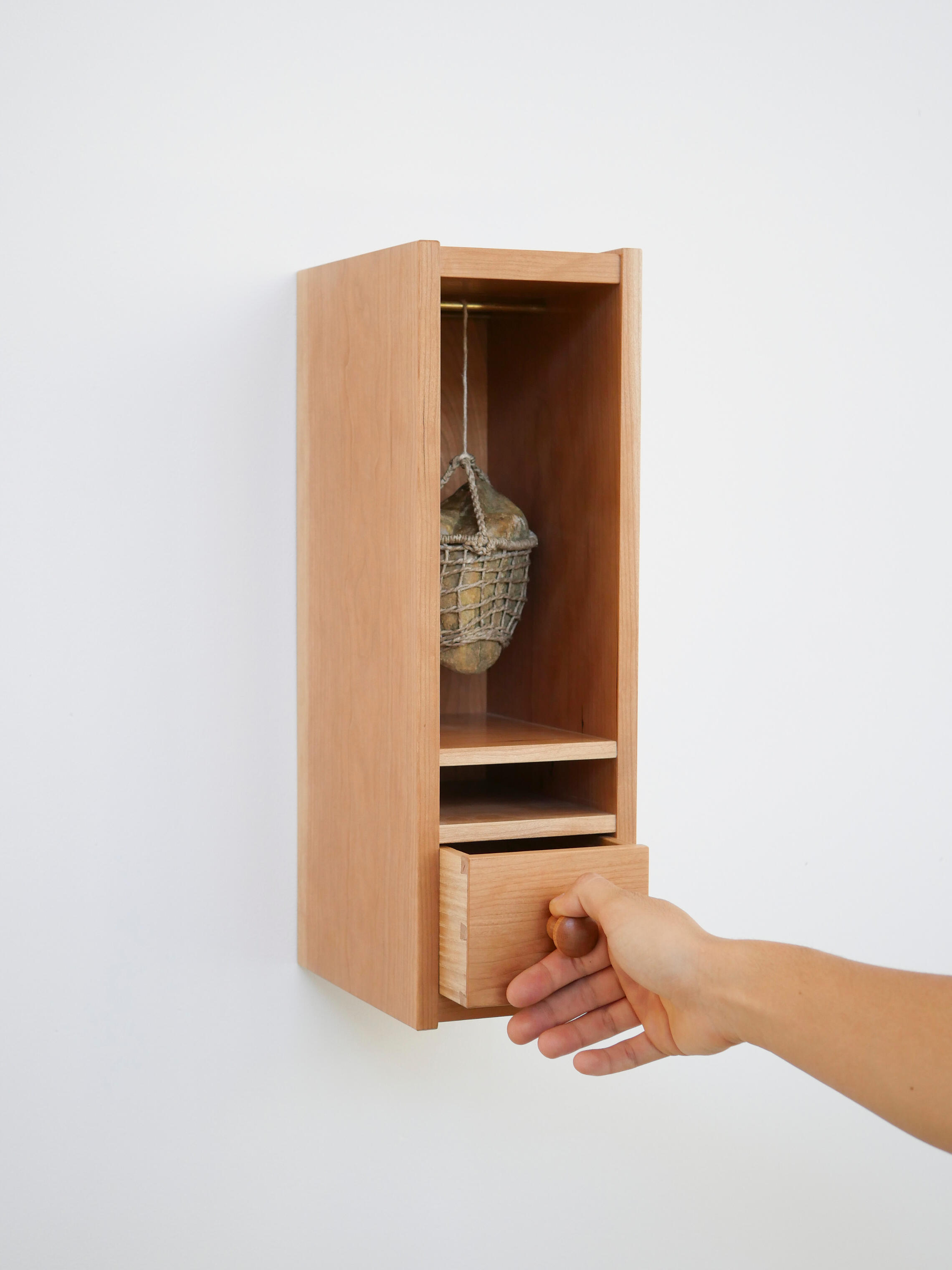 A small cherry wood cabinet hung on a white wall has an open shelf in the top two thirds and a drawer in the bottom one third. The drawer is being partially opened by a hand and that action is lifting a rock on a cable in the upper cavity. Made by Mia Dona